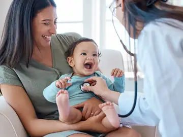 Pediatrician examining a smiling baby on a mother’s lap with a stethoscope at Memorial Heights Emergency Center in Houston, TX